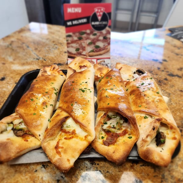Four golden-brown SliceWorks CT calzones filled with cheese and vegetables on a black tray.
