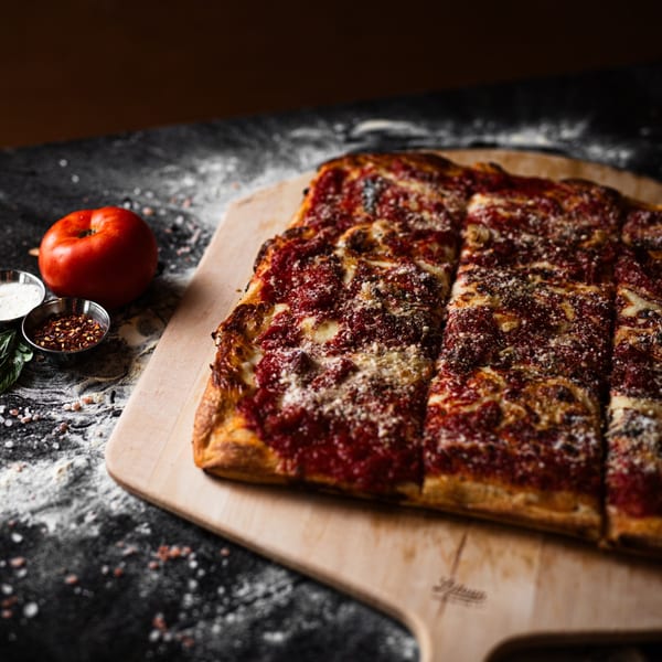 A rectangular SliceWorks CT pizza with red sauce and cheese, on a wooden peel with a tomato and spices.