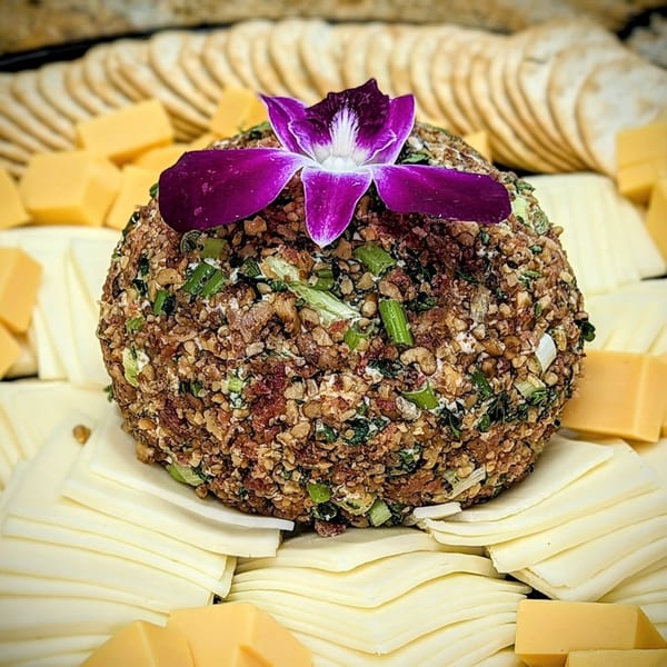 A cheese ball with nuts and green onions, garnished with a purple orchid, surrounded by crackers and cheese.