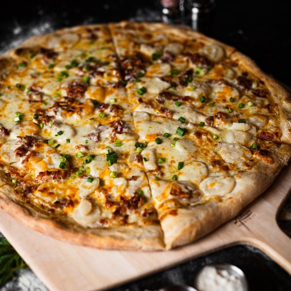 A SliceWorks CT pizza with white and orange cheese, bacon, and chives on a wooden peel.