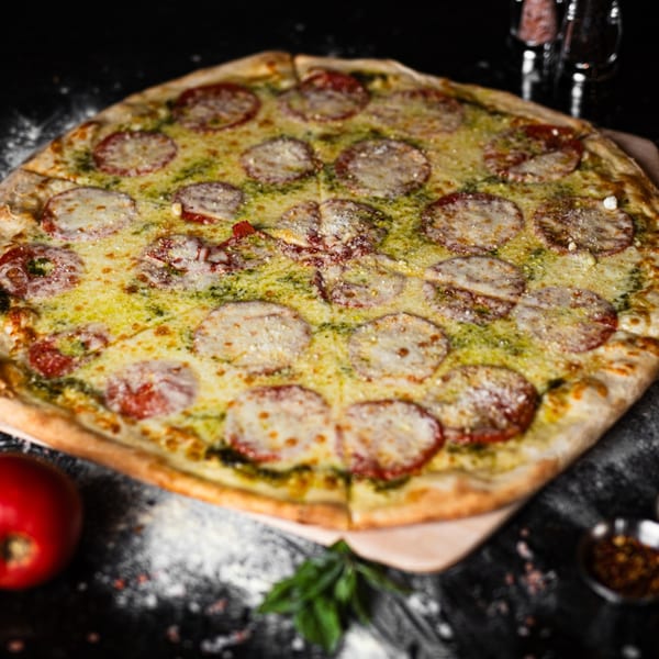 A large, sliced pesto pizza with mozzarella and tomato slices on a dark surface.