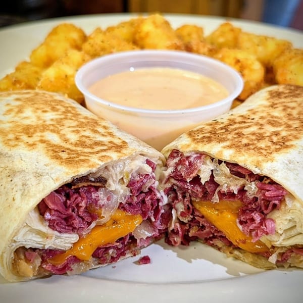 A sliced Reuben wrap with corned beef, cheese, and sauerkraut, served with tater tots and dipping sauce.